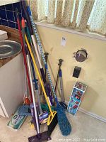 Cleaning tools including multiple brooms, dust mops, and Swiffer mop leaning against a wall near a sink