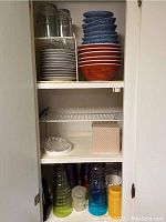 Four shelves inside a white cupboard containing white side plates, patterned orange and blue bowls, empty middle shelf with a clear glass dish and pink polka dot box, and colorful plastic drinking glasses stacked on lower shelf.