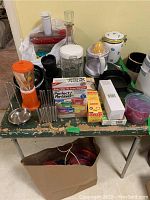 Photo showing the full lot contents on an old green table including blender, knives, decanter, juicer, food storage containers, lidded jar, and other kitchen tools.