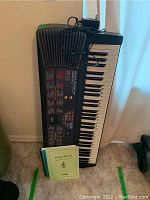 Casio CTK-555LS keyboard leaning against a wall on carpet, with two instruction booklets on the floor beside it and a power adapter resting on top.