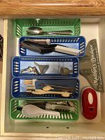 Overhead photo showing trays with cutlery including spoons, forks, can opener, and kitchen tools in plastic trays of green and blue.