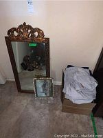 Photo showing ornate framed mirror, smaller framed village art, and stacked bed linens on box.