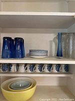 Two shelves of kitchenware showing blue glassware, clear glass vases, stack of small blue-rimmed plates, ceramic cups with blue floral design, and nested vintage Pyrex mixing bowls in yellow, cream, and blue.