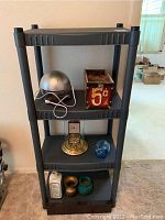 Black plastic shelving unit with assorted items displayed on shelves including a metal air infuser, a 5¢ box, a mantle clock, piggy bank, small ceramic bottle, and decorative pots.
