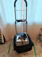 Photo of the collapsible Trolly Dolly hand cart carrying the boxed audio speakers on top, showing the metal frame, padded grip handles, and black fabric bag.