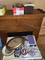 Vintage style oval wall clock with bronze frame at front, surrounded by decorative figurines including dolphin and flower-shaped ceramic containers, remote control, and pill sorter on a white tray.