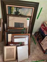 Photo showing stack of framed prints of various sizes and styles leaning against the wall on carpeted floor.