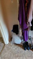 Photo showing various women's clothing items hanging, including a purple garment and patterned items, with black boots on the floor and a plastic bag underneath clothing rack.