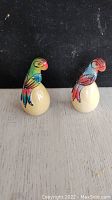 Front view of the two ceramic parrot figurines showing colorful painted details on bodies and egg-shaped bases.