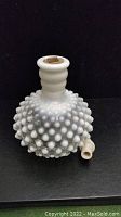Side angle of white hobnail milk glass bottle with stout round body and narrow neck, showing textured bumps and attached glass spout at base