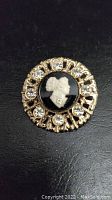 Front view of the vintage cameo brooch showing white woman's profile on black background surrounded by gold-tone metal and clear rhinestones.