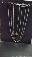 Front view of multiple silver-tone chains hung on black background, featuring a locket pendant on one chain.