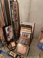 Overview showing approximately 15 framed art prints and posters leaning against wall in basement storage