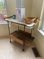Photo showing beverage cart with white top, wood and iron frame, with small wheels, situated in a kitchen corner, some items on top but not included.