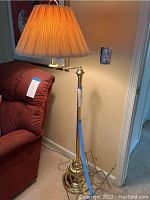 Full view of tall brass swivel standing lamp with pleated cream lampshade beside a red chair, showing the height and lighting features