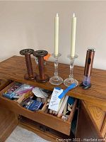 Two wooden candleholders, two crystal-like glass candleholders with tall white taper candles on a wooden surface, drawer below containing matches and other items.