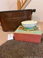 Oblong copper trough bin with dark patina and two decorative hand-painted wooden boxes stacked in front of it on carpet.
