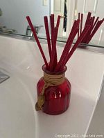 Red glass oil diffuser with red reed sticks and twine, placed on bathroom sink counter in front of a mirror