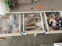 Top view of three open garage drawers containing various homeowner supplies including power strips, plastic droppers, tools, and rope.