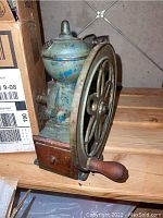 Antique iron coffee grinder with a cast iron body, large wheel, wooden handle, and small wooden drawer at the base on wooden surface.