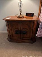 Front view of one oak nightstand with closed double doors and beveled edge top surface. Lamp and clock rest on top.