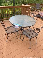Round glass top outdoor table and four metal mesh chairs on wood deck, showing full set and environment.