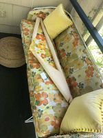 Full view of Homecrest outdoor wrought iron sofa with floral cushions, yellow pom pom throw pillow, and rolled extra fabric on sofa seat. Jute pouf ottoman to the side.