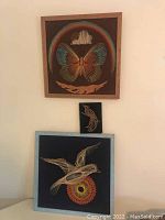 Framed string art featuring a colorful butterfly, small fish, and bird designs on black backgrounds with wooden frames, arranged on a wall and table surface.