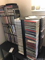 Side view showing two metal wire stands loaded with a mixture of CD and DVD cases tightly packed with various music genres including jazz, classical, opera.