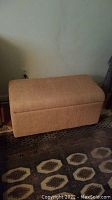 Front view of the closed rectangular beige upholstered storage ottoman on patterned carpet.