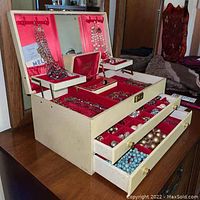 Open cream-colored jewelry chest with red interior showing assortment of bead necklaces and jewelry inside and in drawers.