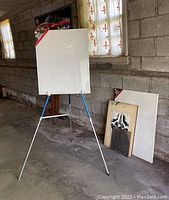 Photo of metal folding easel holding a large stretched canvas in shrink wrap inside a garage with concrete block walls and concrete floor.