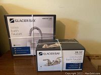 Two Glacier Bay bath faucets in sealed packaging, front views showing brand and model numbers