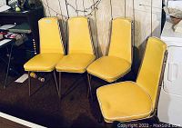 Four yellow vinyl dining chairs with white piping, mid-century modern style, arranged against wood panel wall.