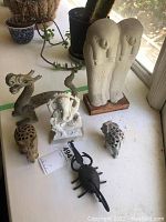 Seven decorative figurines arranged on a windowsill, including a dragon, elephant head, Ganesha deity, scarab, scorpion, and Egyptian mummy figures.
