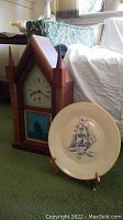 Wooden nautical wall clock with pointed arch and two finials, clock face with Roman numerals, lower panel showing ship painting. Next to it is a Lenox display plate on a stand showing a sailing ship.
