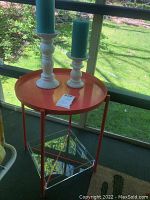 Full view of round orange metal table with removable top and clear plexiglass lower tray. Two white candlestick holders with blue pillar candles placed on top. Background shows screened porch and garden.