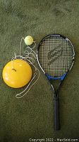 Full view of Slazenger tennis racket lying on a green carpet next to tethered training ball and yellow base.