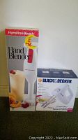 Shows Hamilton Beach hand blender and Black & Decker hand mixer in their original retail boxes placed side by side on a floor surface.