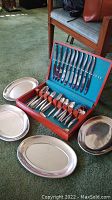 Wooden box flatware set with multiple utensils inside, placed on green carpeted floor with 4 oval stainless steel platters arranged around it