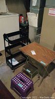 Photo showing black metal wheeled cart, wood-patterned snack table, purple folding stool, and green step stool in basement setting