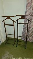 Two wooden vintage gentleman's butler stands with wheels, standing side by side against a corner wall. Both have horizontal bars for hanging, hanger-shaped frame, and wheels at the base.