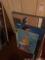 Photo of a group of original paintings and prints stacked upright against a wall, showing various abstract and figurative artworks with different frames and color schemes.