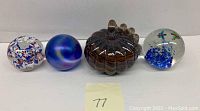 Front view of four art glass paperweights in a row on white surface: floral confetti design, iridescent blue swirl, pumpkin shaped brown glass, clear glass with blue bubbles and red fish.