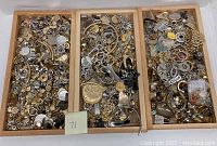 Three wooden trays filled with an assortment of loose costume jewelry pieces. Items include various earrings, chains, pendants, and rings in gold and silver tones with some decorative details.