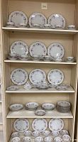 Full view of shelves displaying multiple sets of dinner plates, salad plates, bread plates, cups, saucers, bowls, creamer, and sugar bowl, all with floral patterns and gold trim.