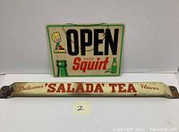 Photo showing both signs: rectangular Squirt open sign with a bottle and character and curved Salada Tea porcelain door push sign with red text.