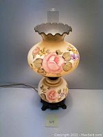 Front view of vintage floral hurricane lamp showing the painted pink roses and yellow and purple flowers on frosted glass, with ruffled top and clear glass chimney visible.