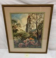 Framed watercolor painting showing a church tower amidst trees and blooming garden flowers with a beige mat and wooden frame.