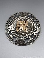 Front view of round brooch showing central gold embossed motif on textured silver background with open cutout animal and geometric designs surrounding it.